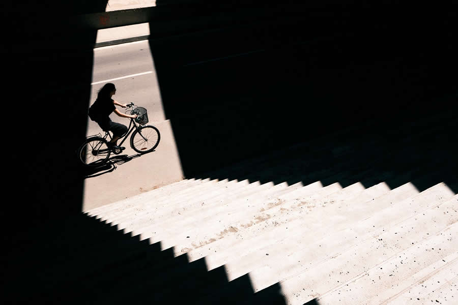 The Art of Perfect Timing: 34 Clever Street Photos by Emiliano Cuadrado That Turn Ordinary Scenes Into Visual Surprises 39 Silhouette of a cyclist riding through a narrow shaft of sunlight beneath an overpass, framed by deep shadows and geometric staircase patterns in a minimalist street photography composition.
