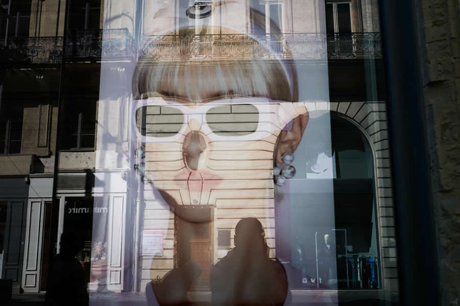 The Art of Perfect Timing: 34 Clever Street Photos by Emiliano Cuadrado That Turn Ordinary Scenes Into Visual Surprises 54 Reflection of a fashion advertisement featuring a woman in white sunglasses layered with historic building facades and a silhouetted pedestrian, creating an abstract, multi-layered street photography composition.