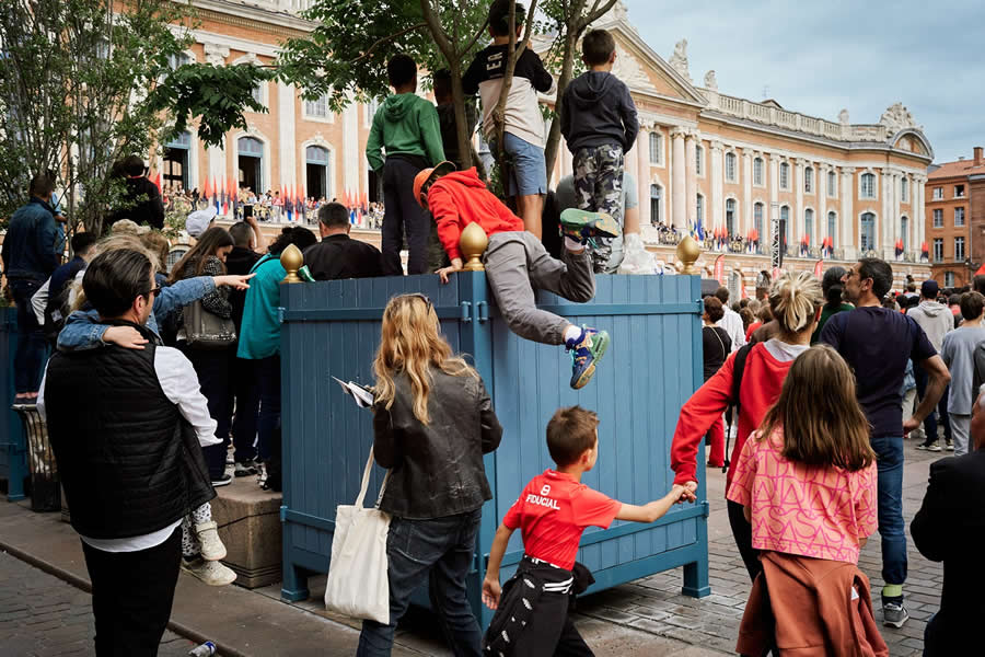 The Art of Perfect Timing: 34 Clever Street Photos by Emiliano Cuadrado That Turn Ordinary Scenes Into Visual Surprises 55 Children climbing onto a blue fence to watch a public event as a crowd gathers in front of a historic building, creating a layered and energetic street photography composition.