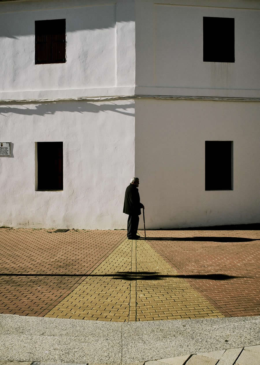 The Art of Perfect Timing: 34 Clever Street Photos by Emiliano Cuadrado That Turn Ordinary Scenes Into Visual Surprises 57 An elderly person with a walking cane stands before a minimalist white building, framed by geometric windows, long shadows, and expansive negative space in a contemplative street photography composition.