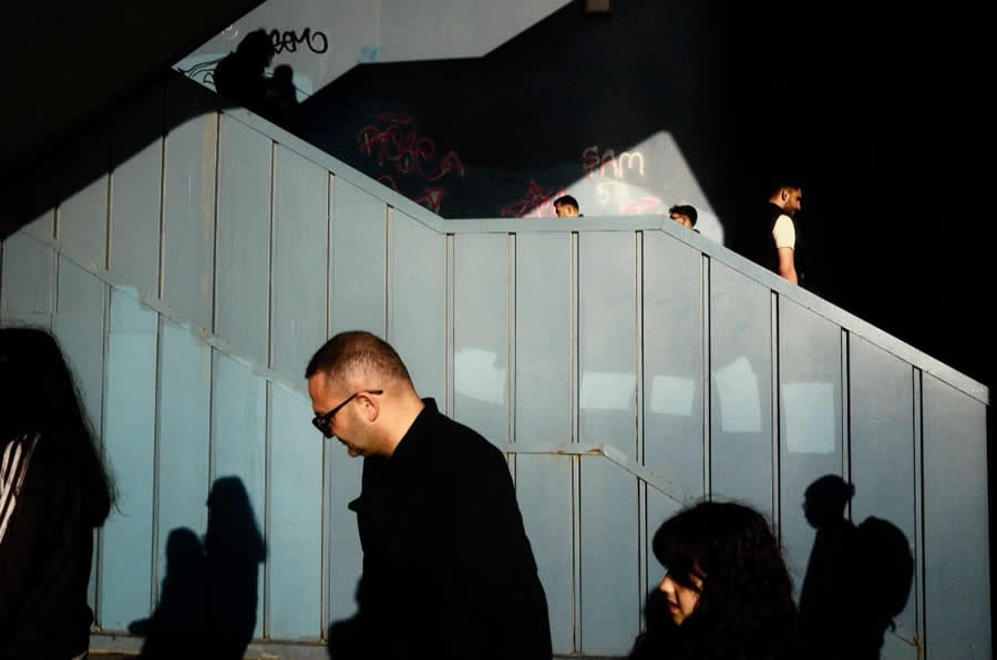 The Art of Perfect Timing: 34 Clever Street Photos by Emiliano Cuadrado That Turn Ordinary Scenes Into Visual Surprises 62 People walking on multiple levels of a modern staircase are framed by strong diagonal lines, dramatic sunlight, and deep shadows, creating a layered geometric street photography composition.