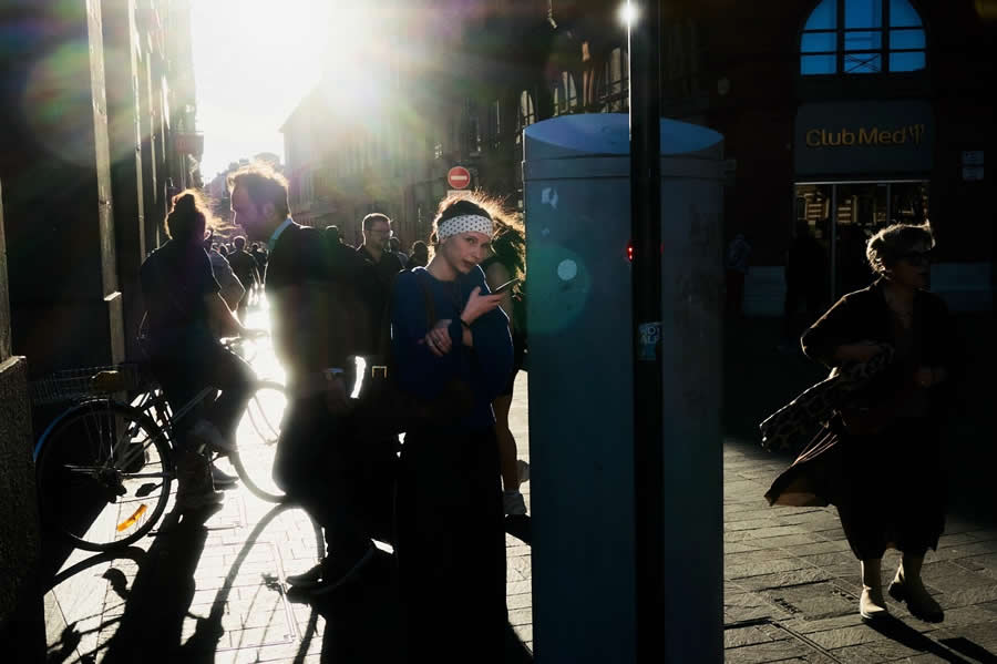 The Art of Perfect Timing: 34 Clever Street Photos by Emiliano Cuadrado That Turn Ordinary Scenes Into Visual Surprises 69 Backlit pedestrians walk through a city street at sunset, with one person pausing near a utility box as dramatic sunlight, silhouettes, and lens flare create a cinematic street photography composition.