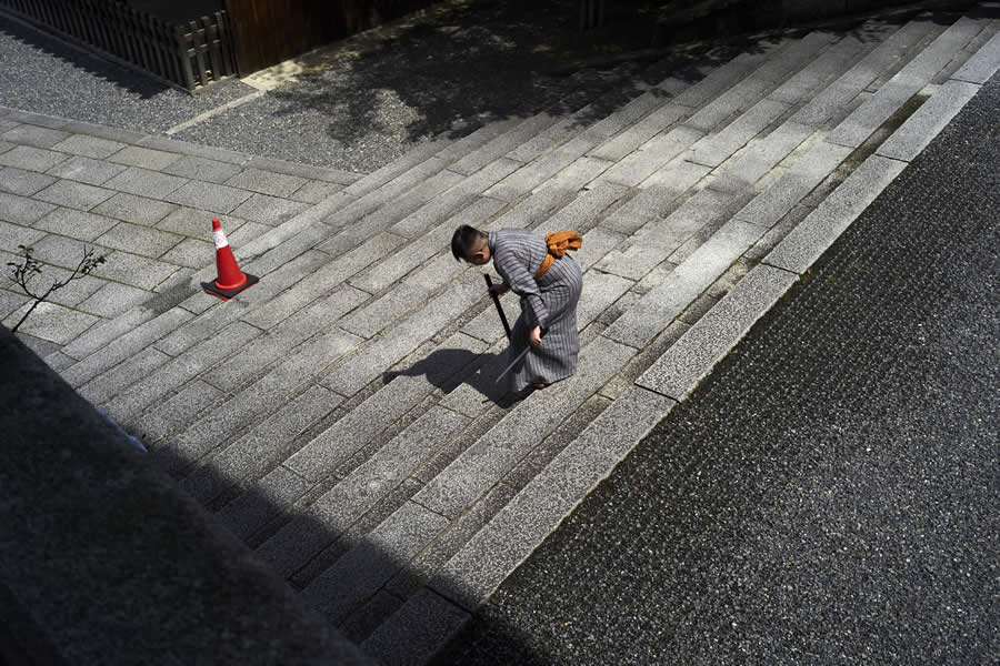 The Art of Perfect Timing: 34 Clever Street Photos by Emiliano Cuadrado That Turn Ordinary Scenes Into Visual Surprises 42 Overhead view of a person in traditional clothing walking up wide stone steps, framed by geometric lines, soft shadows, and a lone orange traffic cone in a minimalist street photography scene.
