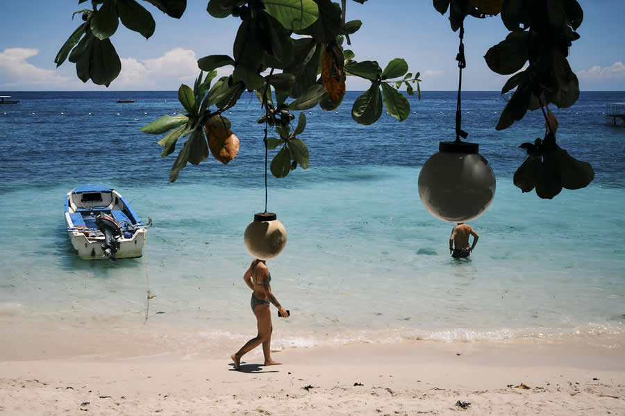 The Art of Perfect Timing: 34 Clever Street Photos by Emiliano Cuadrado That Turn Ordinary Scenes Into Visual Surprises 43 People walking and standing on a tropical beach appear to have hanging lamps instead of heads, framed by tree branches, turquoise water, and a small boat in a perfectly timed street photograph.