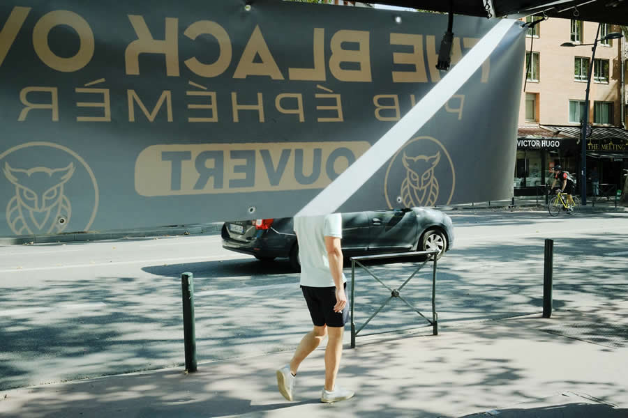 The Art of Perfect Timing: 34 Clever Street Photos by Emiliano Cuadrado That Turn Ordinary Scenes Into Visual Surprises 44 A pedestrian walking behind a large hanging banner appears partially hidden, creating an optical illusion enhanced by bold typography, diagonal sunlight, and clean urban street composition.