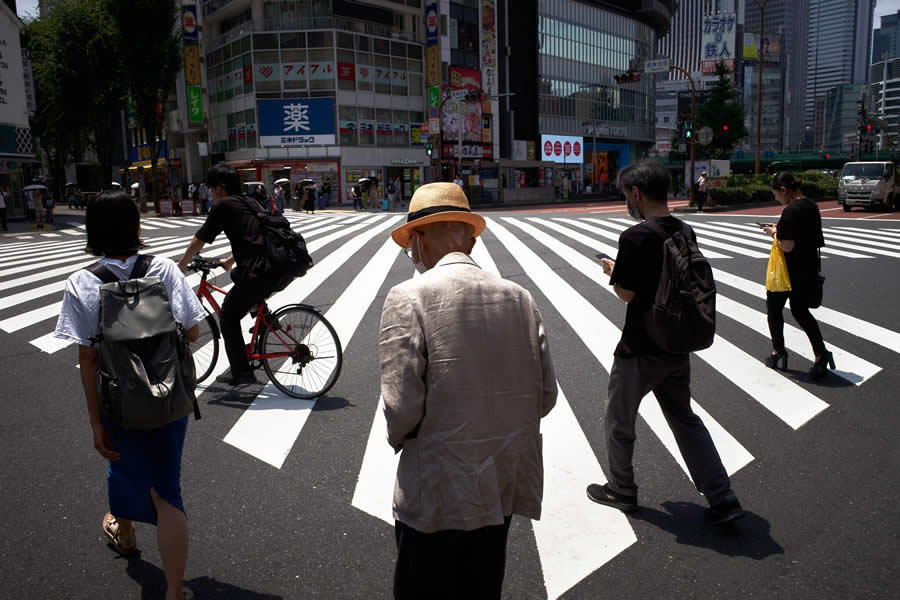 The Art of Perfect Timing: 34 Clever Street Photos by Emiliano Cuadrado That Turn Ordinary Scenes Into Visual Surprises 46 An elderly man wearing a straw hat stands at a wide zebra crossing surrounded by pedestrians and a cyclist, framed by bold geometric lines and a bustling urban cityscape.